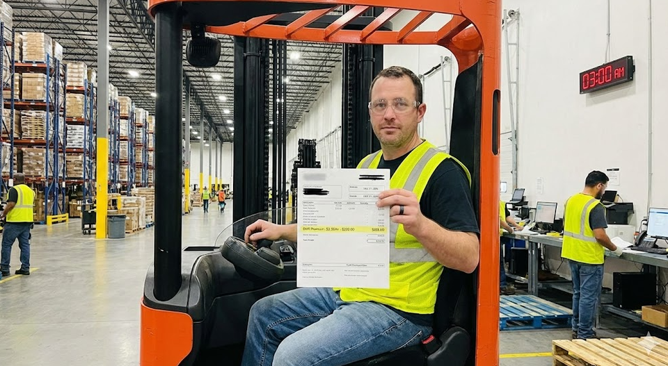 A forklift operator working under bright warehouse lights at 3:00 AM, holding a pay stub showing a "Shift Premium" line item.